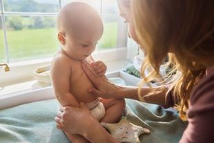Mutter cremt ihr sitzendes Baby am Oberkörper sanft ein – liebevoller Pflegemoment am hellen Fenster.