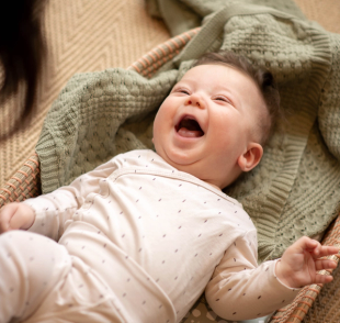 Smiling 6-week-old baby lying on a soft blanket, laughing up at a caregiver during playtime.