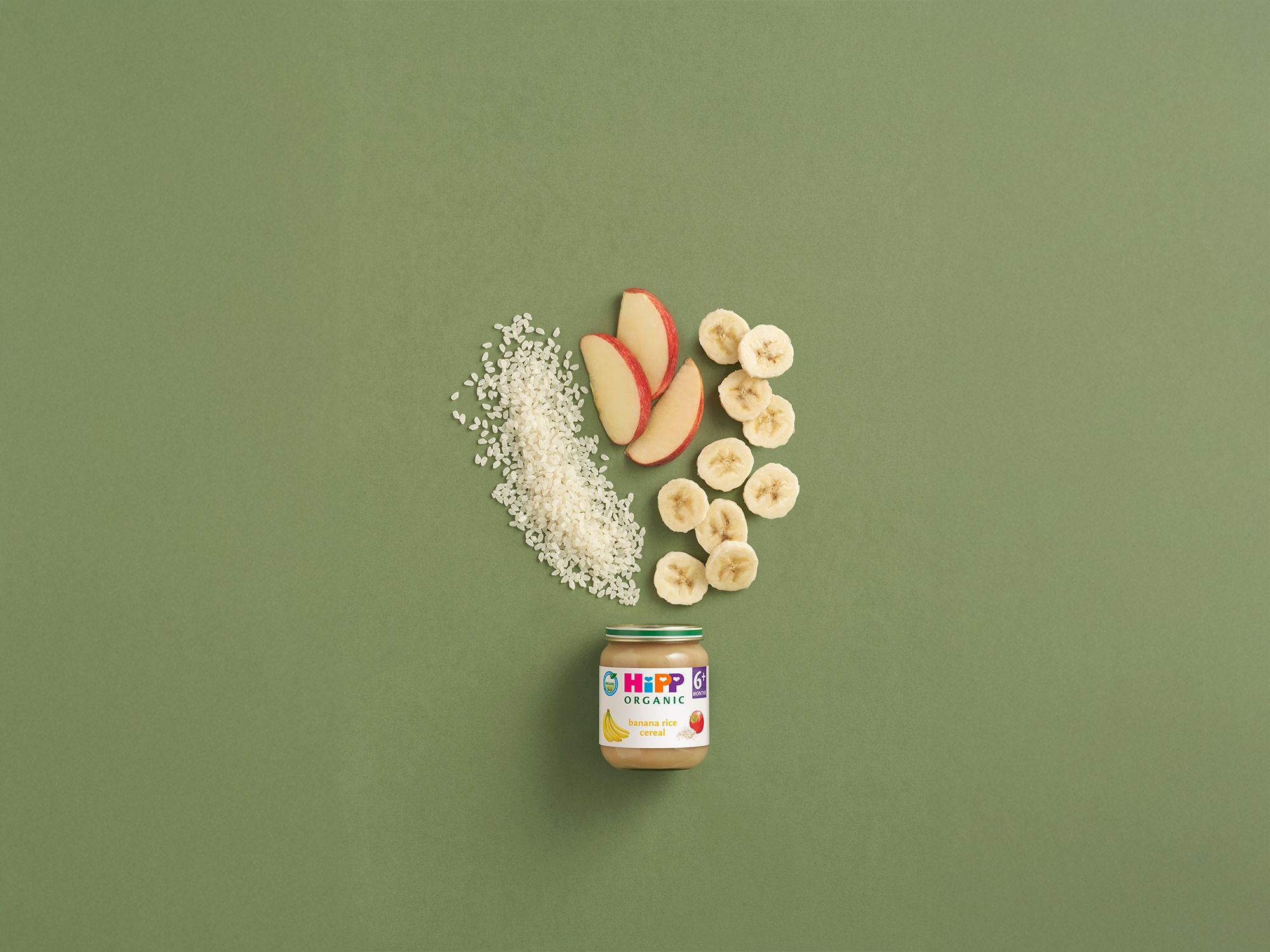 HiPP Organic Banana Rice Cereal jar with rice, banana slices and apple pieces arranged on a green background
