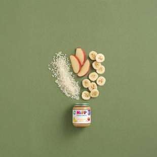 HiPP Organic Banana Rice Cereal jar with rice, banana slices and apple pieces arranged on a green background