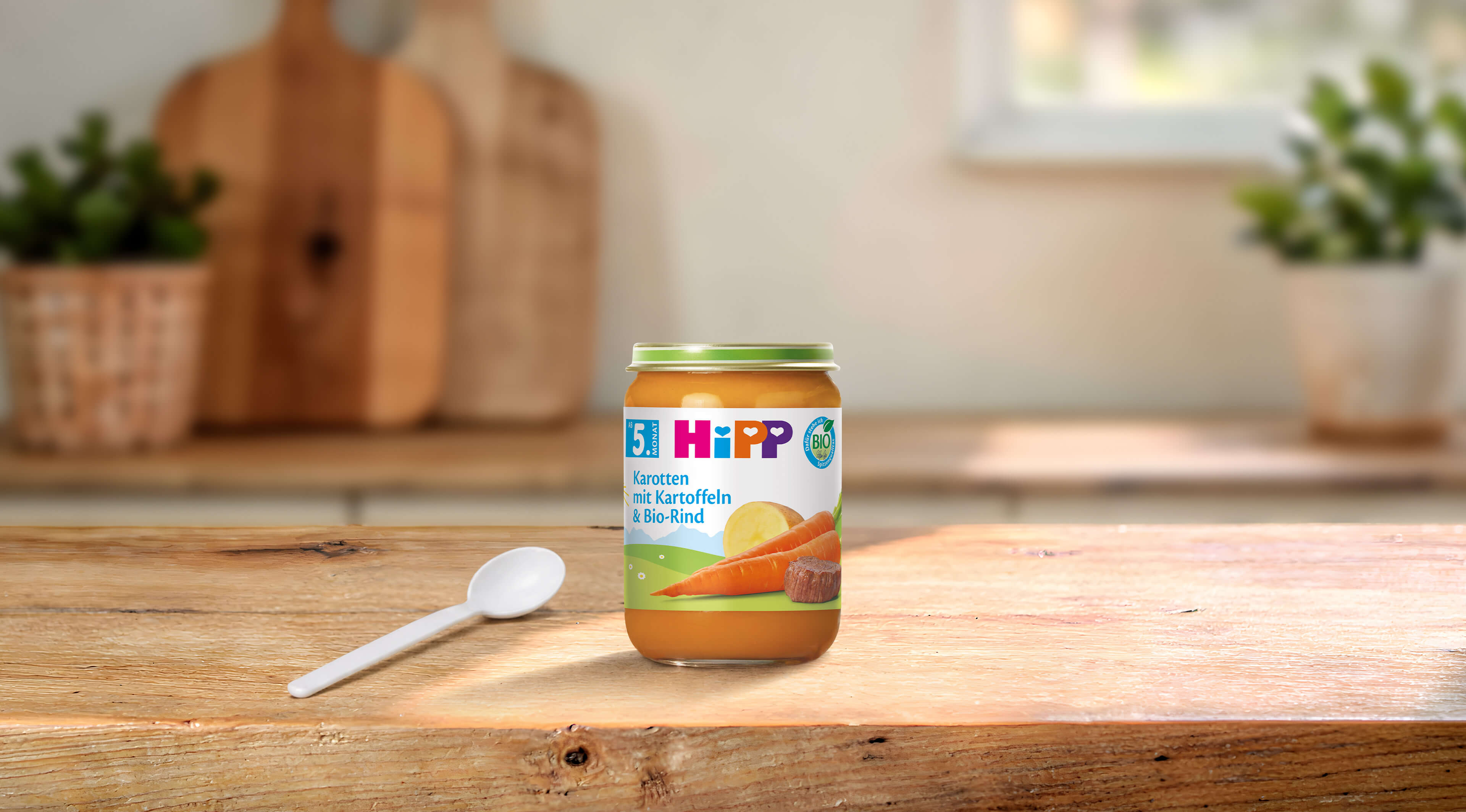 HiPP Bio-Gläschen Karotten mit Kartoffeln und Bio-Rind ab dem 5. Monat auf Holztisch mit Löffel in heller Küche