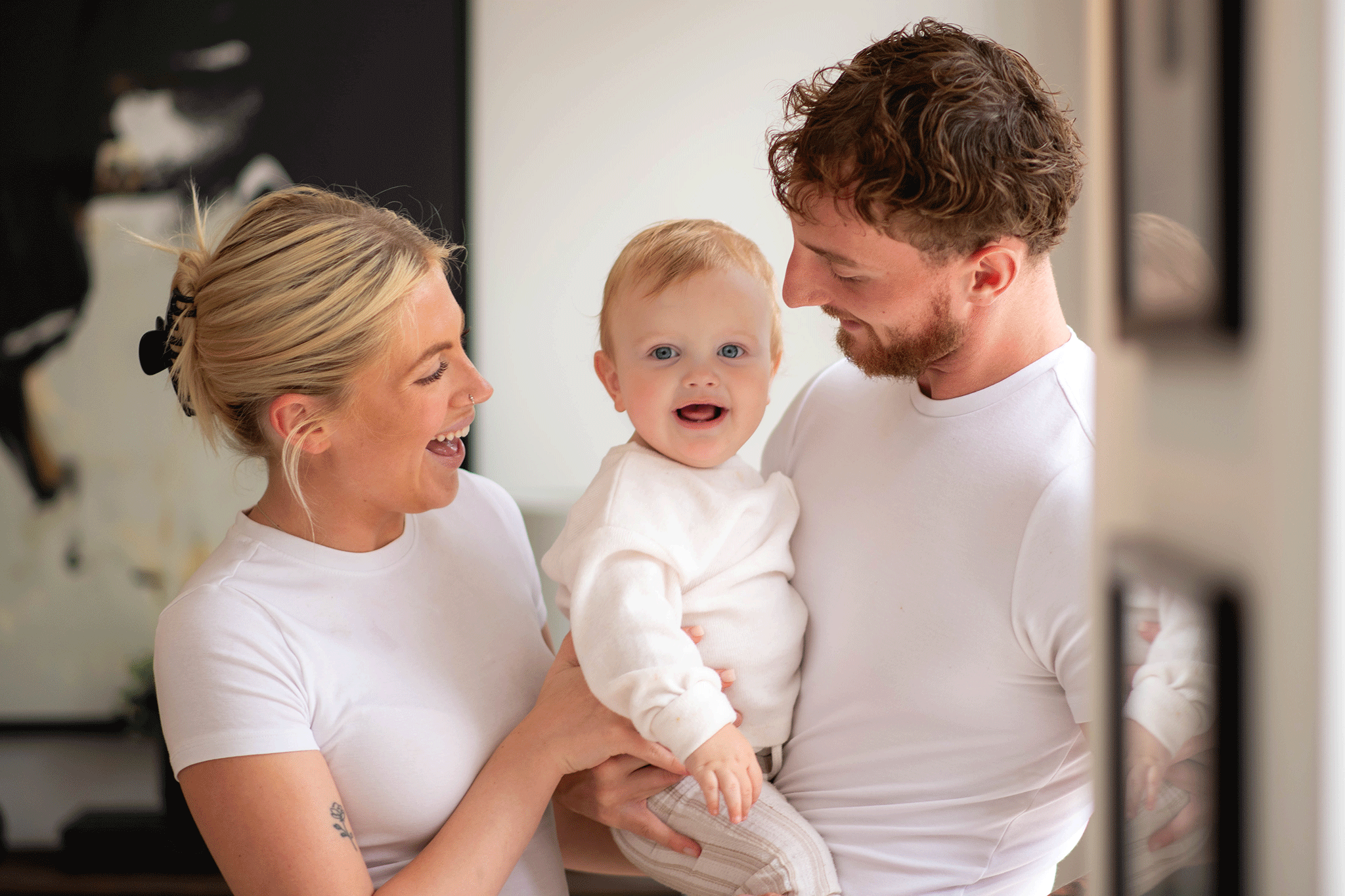 A smiling family portrait featuring a mother and father holding their blue-eyed baby between them, all dressed in matching white t-shirts. The baby looks directly at the camera with an open-mouthed expression, while both parents look lovingly at the child.