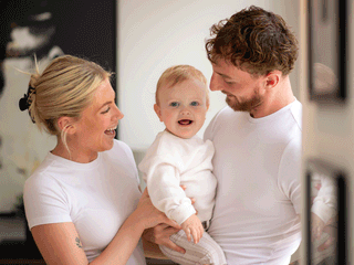 A smiling family portrait featuring a mother and father holding their blue-eyed baby between them, all dressed in matching white t-shirts. The baby looks directly at the camera with an open-mouthed expression, while both parents look lovingly at the child.