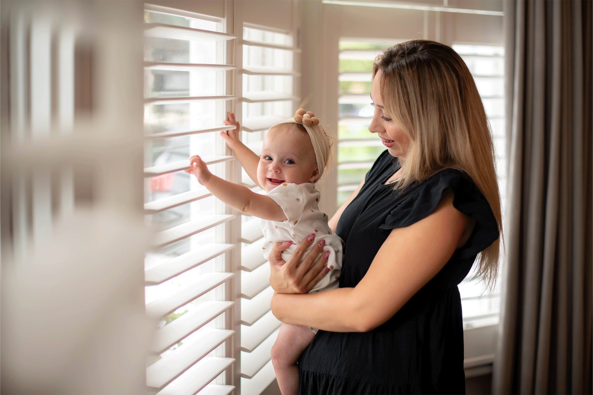 A smiling mother with blonde hair wearing a black dress holds her baby girl near a bright window. The baby, wearing a cream patterned bodysuit and a bow headband, looks back toward the camera while resting her hands on white window shutter slats.