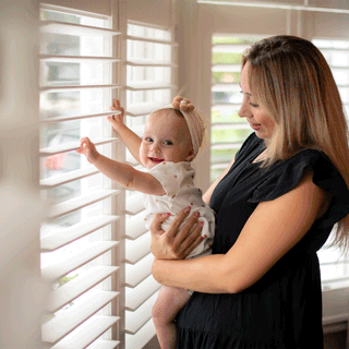 A smiling mother with blonde hair wearing a black dress holds her baby girl near a bright window. The baby, wearing a cream patterned bodysuit and a bow headband, looks back toward the camera while resting her hands on white window shutter slats.