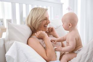 Ein freudiger Moment zwischen einer Mutter und ihrem Baby, wo die Mutter lacht und mit dem Baby auf einem Bett spielt.