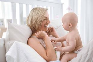 Ein freudiger Moment zwischen einer Mutter und ihrem Baby, wo die Mutter lacht und mit dem Baby auf einem Bett spielt.