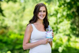 Eine Frau mit einer Wasserflasche in der Hand, die im Freien steht, mit Grünzeug im Hintergrund.