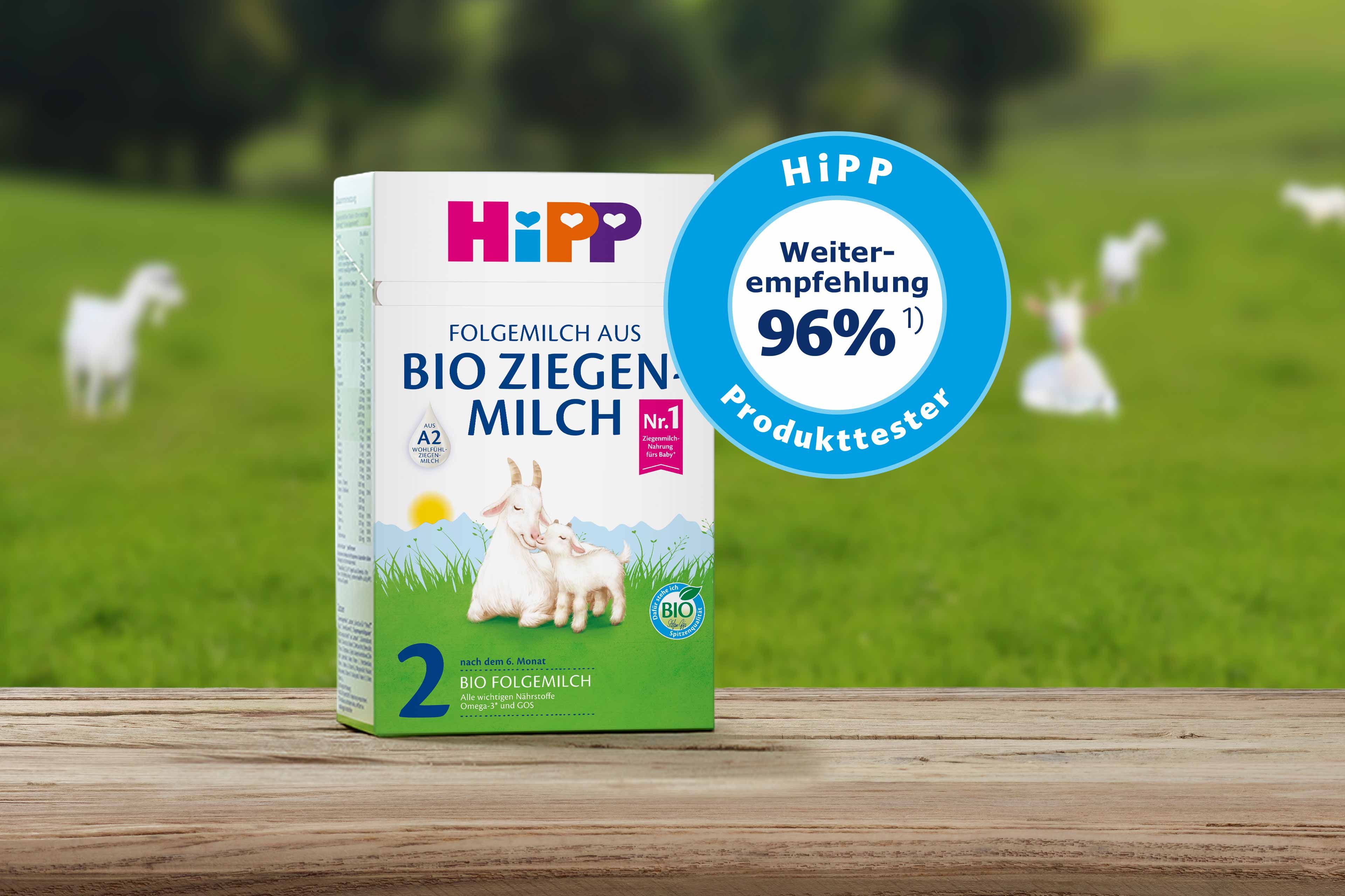 Packung Bio-Ziegenmilch von HiPP mit Ziegenbaby auf Holzuntergrund vor grüner Wiese mit Ziegen im Hintergrund