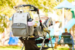 Ein Kinderwagen mit einem Rucksack, der ein Fläschchen und eine Babydecke enthält, vor der Kulisse eines Spielplatzes.