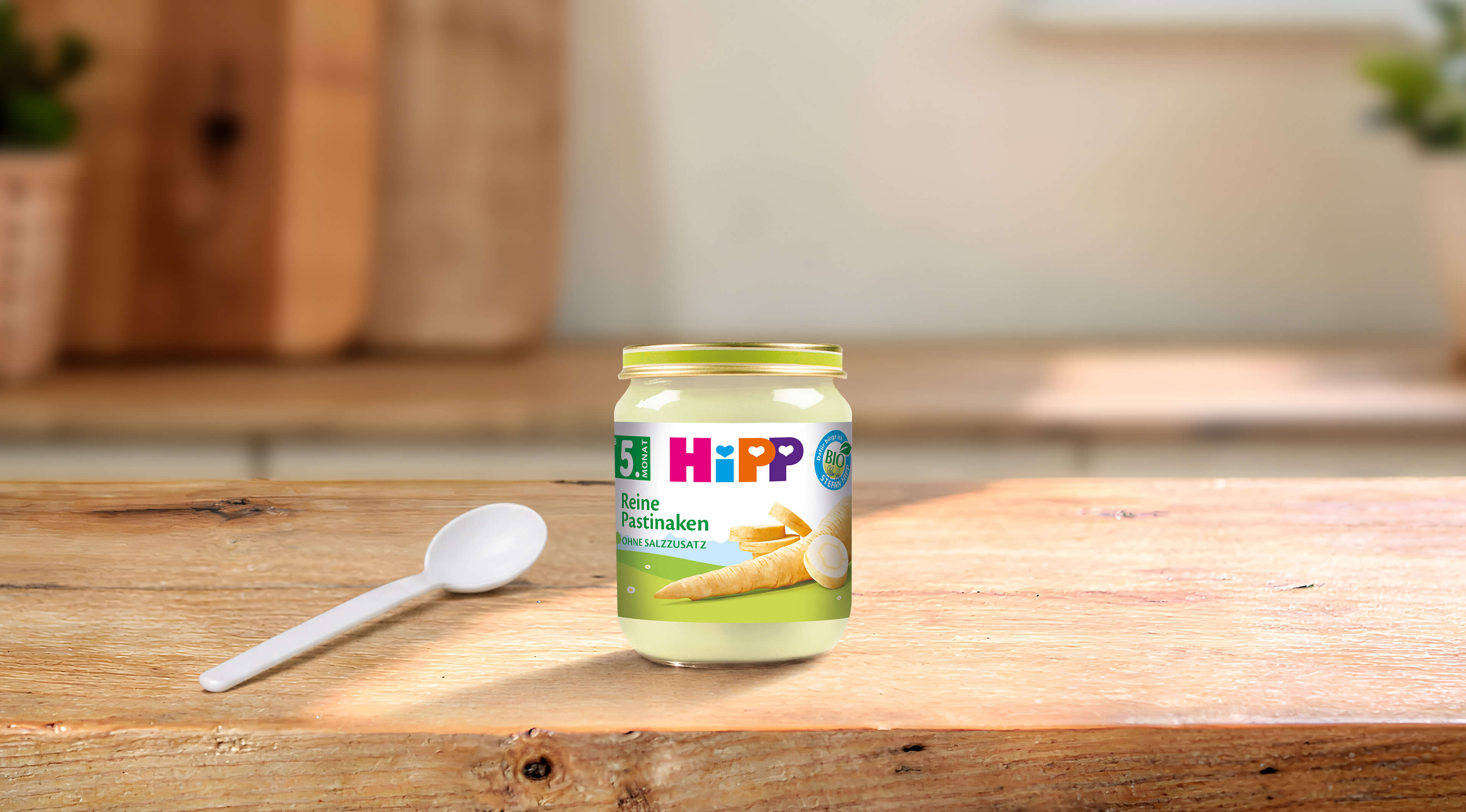 HiPP Bio-Gläschen „Reine Pastinaken“ ab dem 5. Monat mit Löffel auf einem Holztisch in heller Küchenatmosphäre