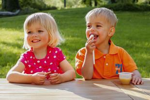 Zwei kleine Kinder, ein Mädchen und ein Junge, sitzen an einem Tisch im Freien, wobei der Junge etwas isst und das Mädchen einen kleinen Gegenstand hält.