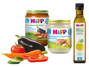 Eine Reihe von Babynahrungsprodukten der Marke "Hipp", begleitet von frischem Gemüse wie Tomaten, Karotten und Gurken.
