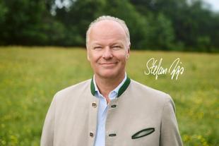 Porträt eines freundlich lächelnden Mannes in traditioneller Jacke vor grünem Hintergrund, daneben seine handschriftliche Unterschrift.