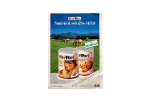 Historische HiPP-Anzeige für Bio-Säuglingsmilch mit zwei Milchdosen im Vordergrund und grasenden Kühen auf grüner Almwiese.