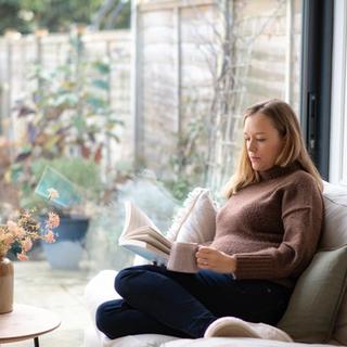 Woman relaxing on a couch by a large window, holding a mug and reading a book, with a vase of flowers and candles on a nearby table.