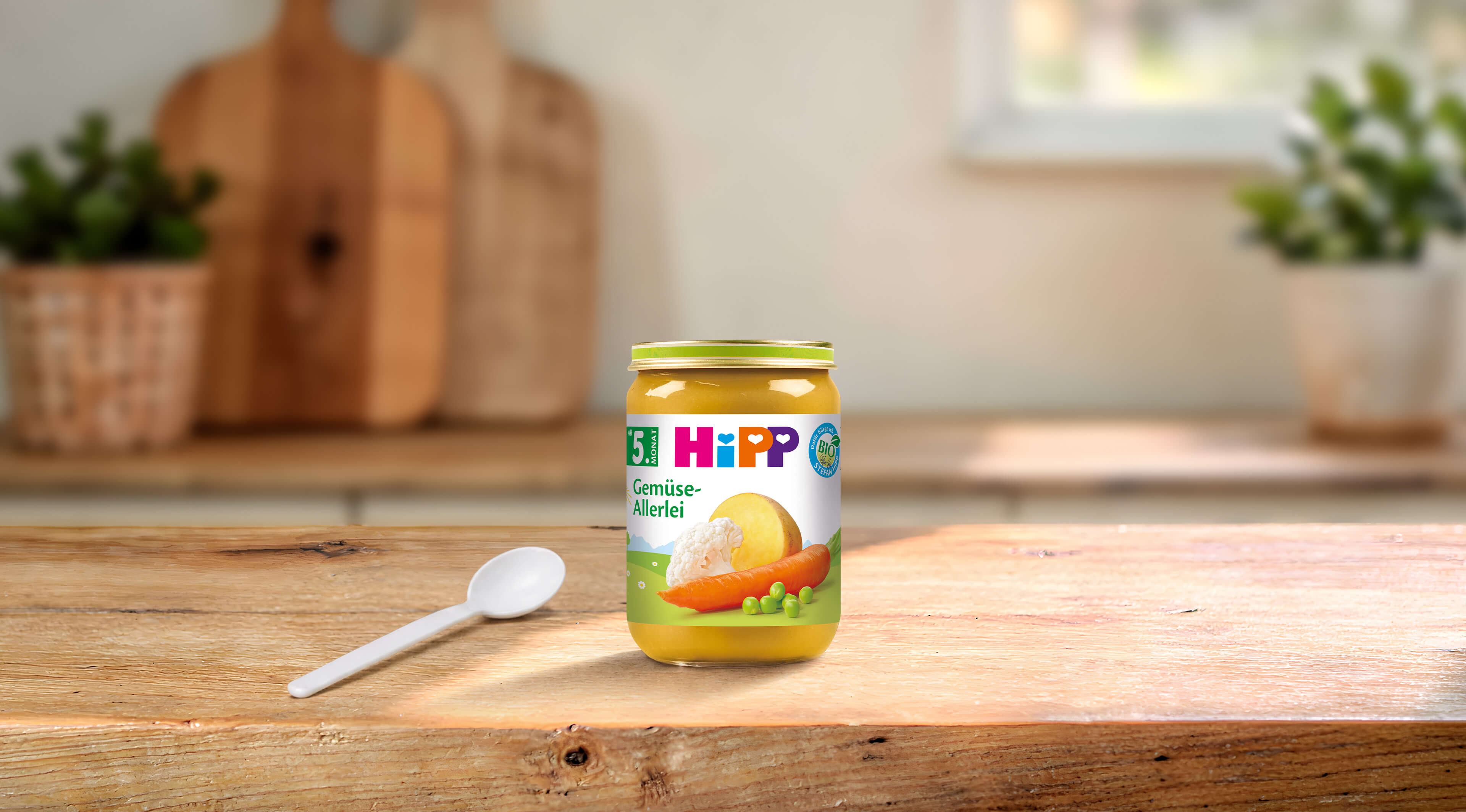 HiPP Gemüse-Allerlei Bio-Gemüsebrei im Glas auf Holztisch mit Löffel, im Hintergrund eine helle Küche