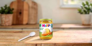 HiPP Gemüse-Allerlei Bio-Gemüsebrei im Glas auf Holztisch mit Löffel, im Hintergrund eine helle Küche