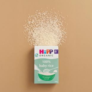 HiPP Organic 100% baby rice box on a neutral background with baby rice grains scattered above it.