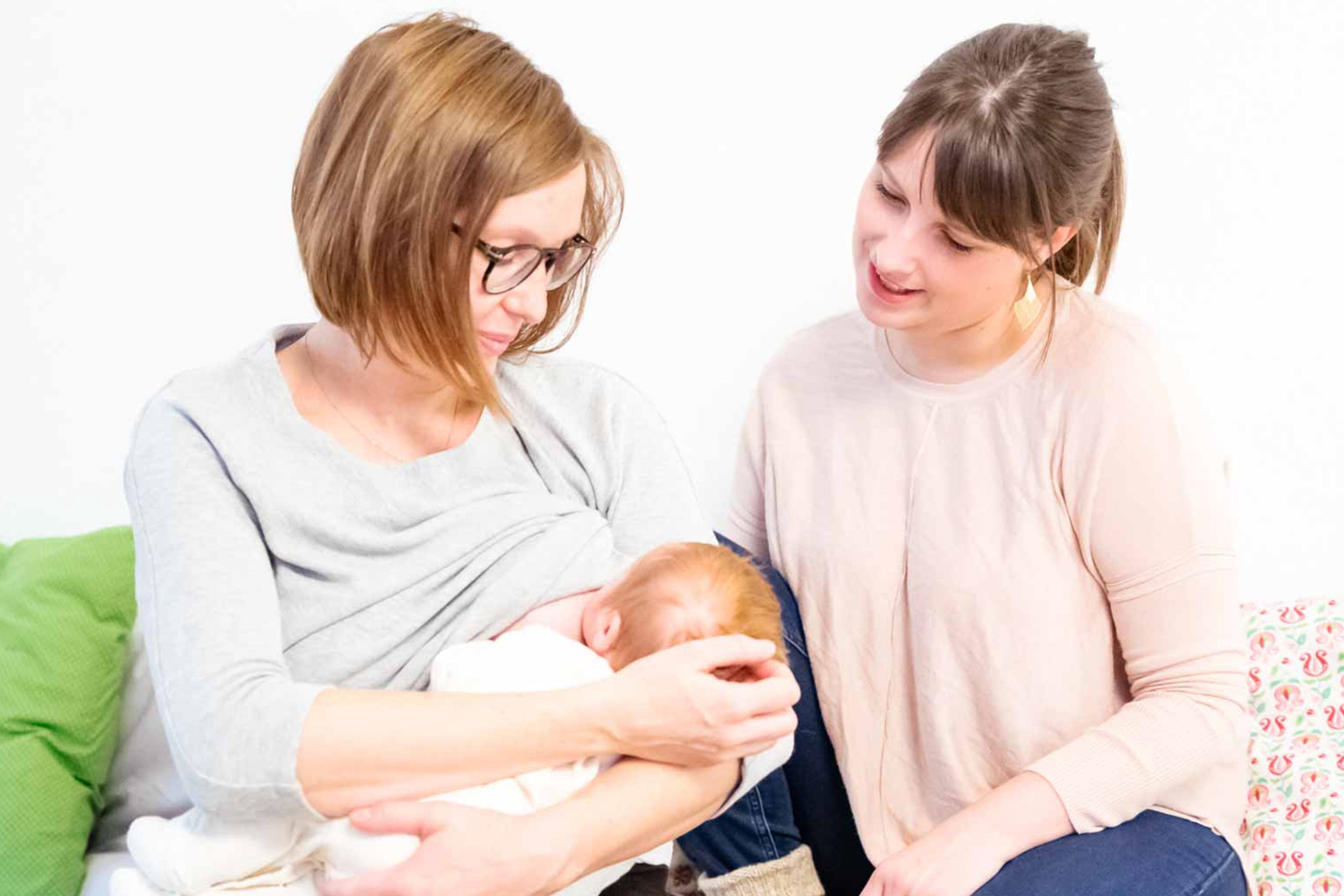 	Zwei Frauen, eine mit einem Baby in der Hand, in einem Raum mit weißem Hintergrund und bunten Kissen.
