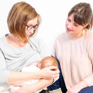 Zwei Frauen, eine mit einem Baby in der Hand, in einem Raum mit weißem Hintergrund und bunten Kissen.
