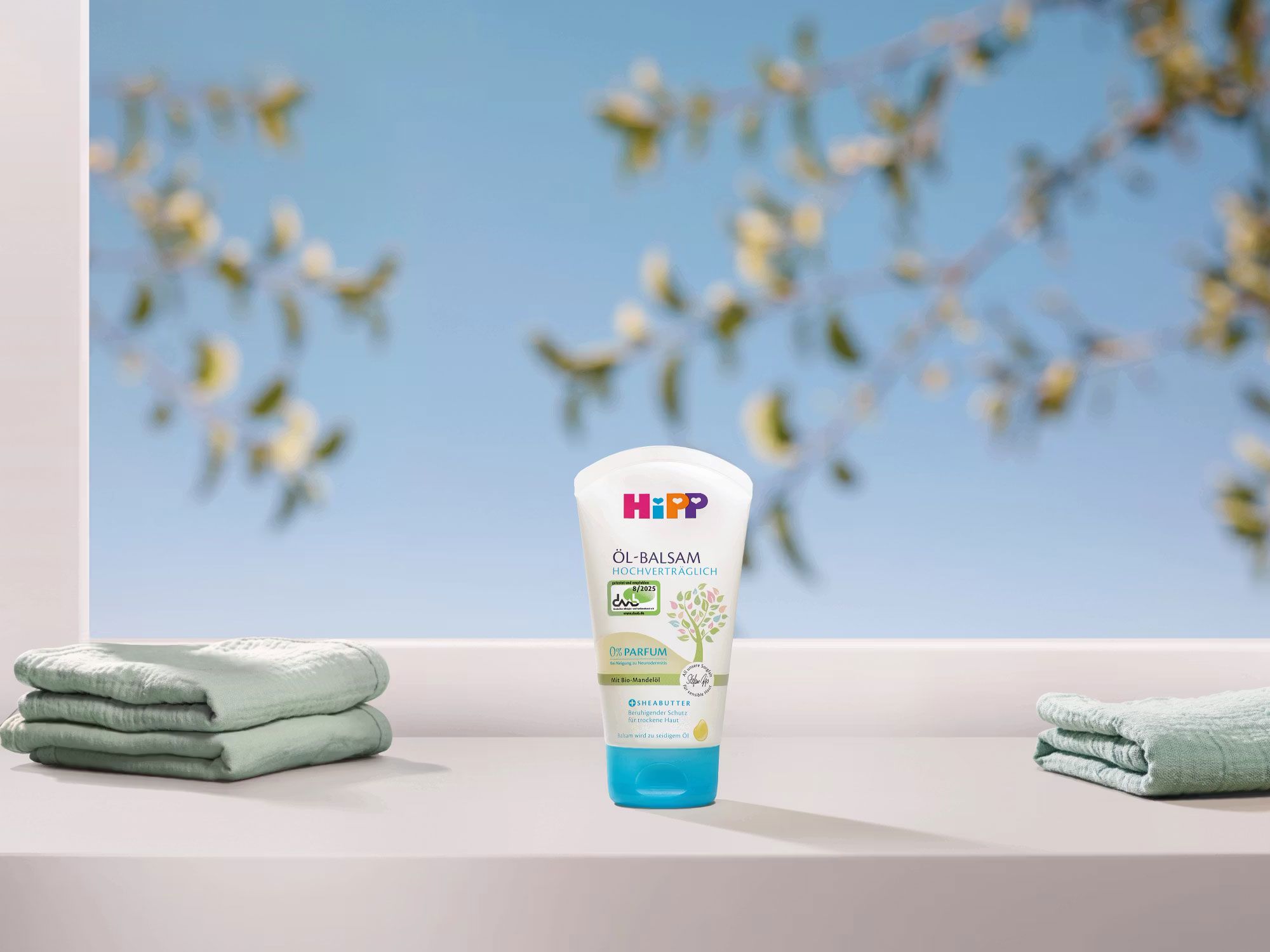 HiPP HOCHVERTRÄGLICH Öl-Balsam Tube vor blauem Himmel, flankiert von weichen Babyhandtüchern, für trockene, empfindliche Babyhaut