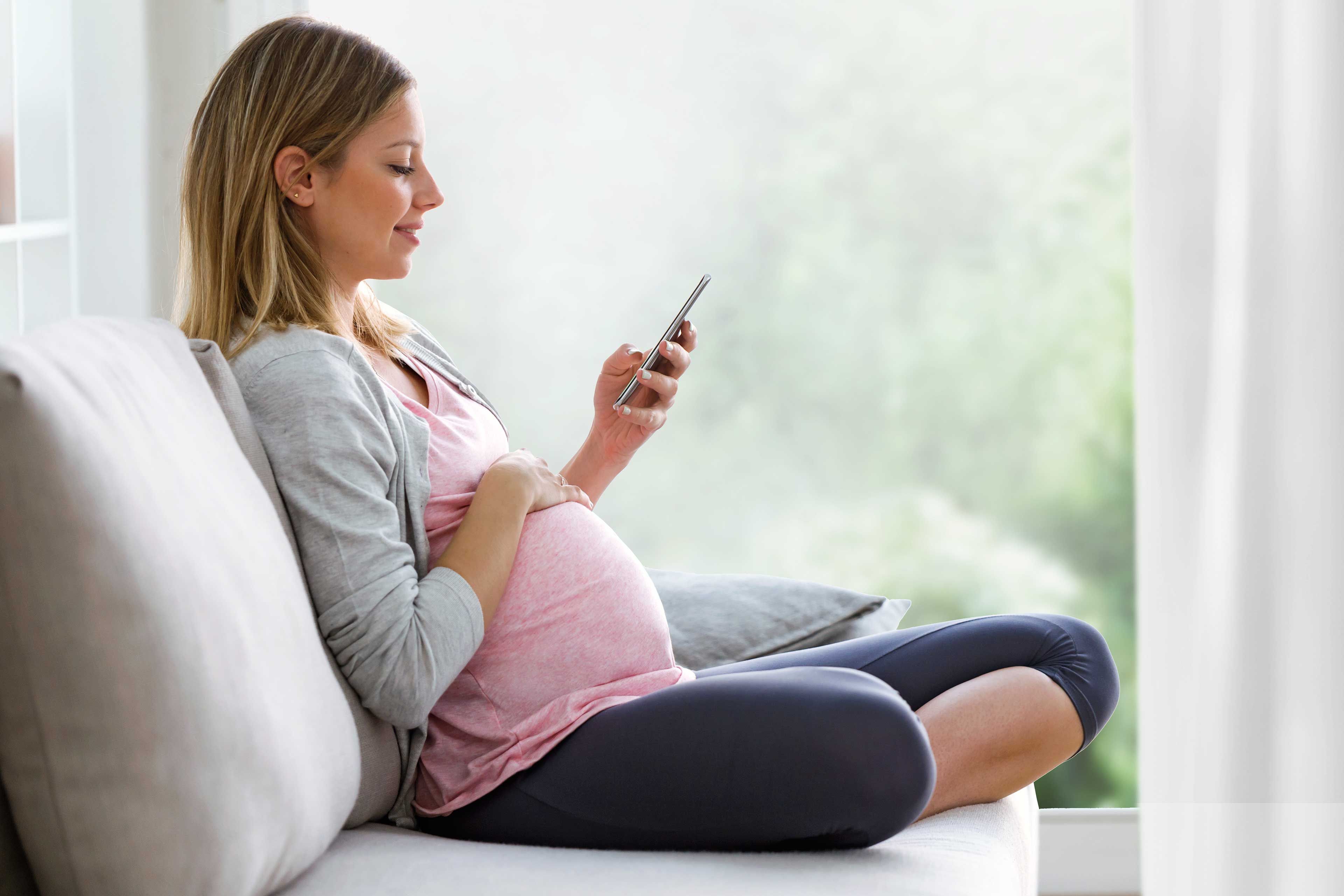 Schwangere Frau sitzt entspannt auf dem Sofa und nutzt auf ihrem Smartphone den HiPP Rechner.