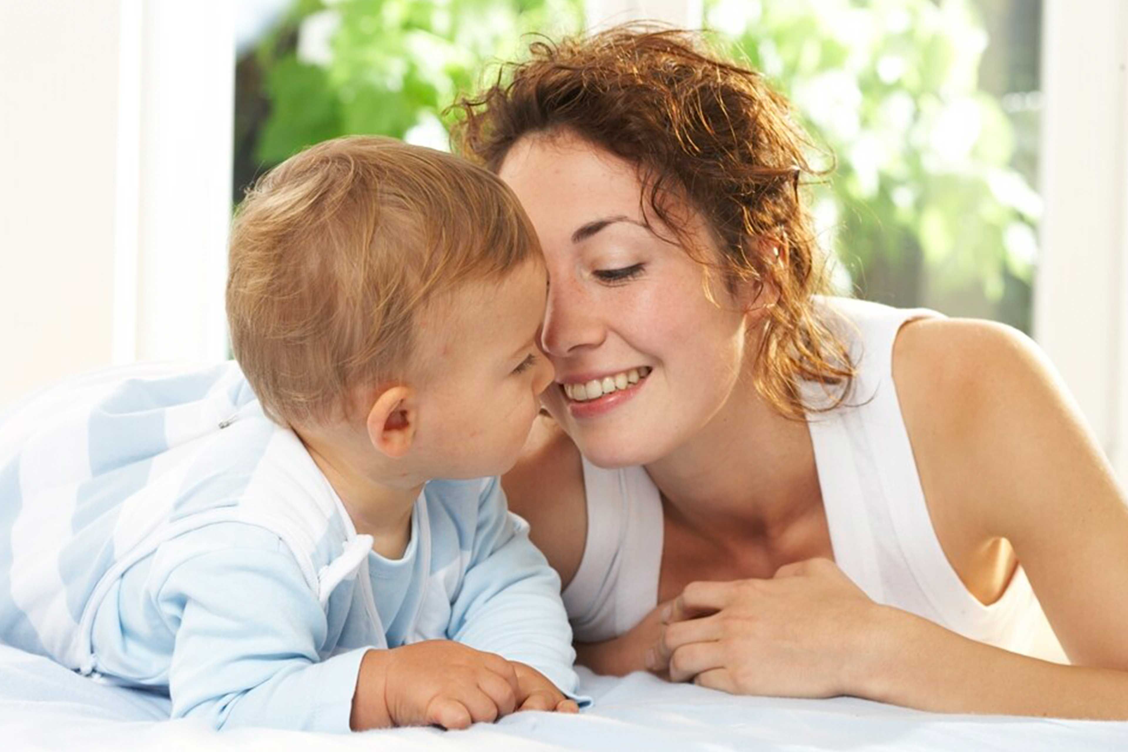Mutter liegt mit ihrem Baby Bauch an Bauch im Bett und berührt liebevoll die Nasen, beide wirken zufrieden und geborgen.