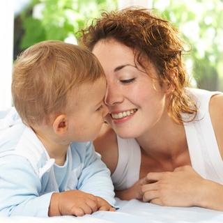 Mutter liegt mit ihrem Baby Bauch an Bauch im Bett und berührt liebevoll die Nasen, beide wirken zufrieden und geborgen.