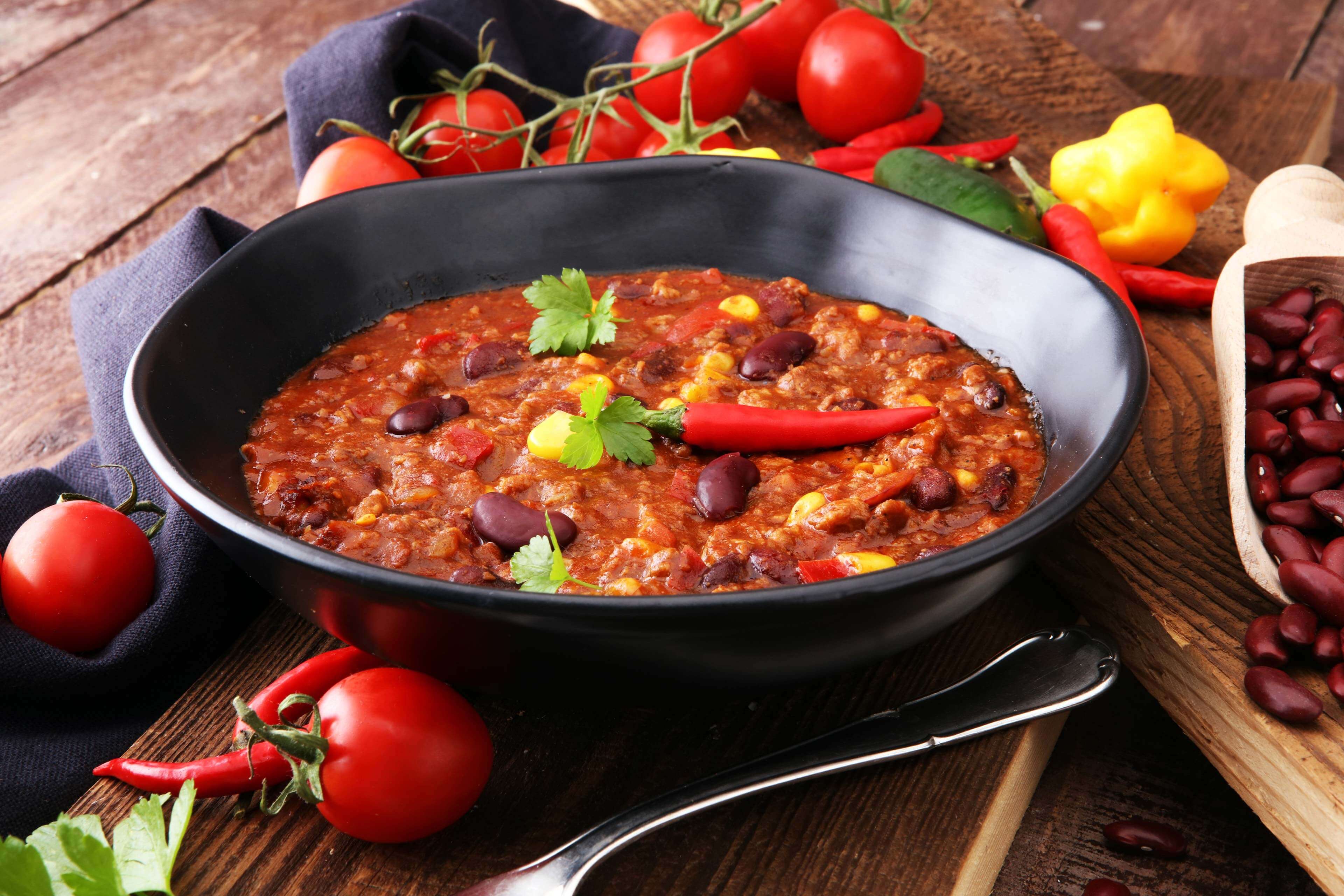 Eine Schüssel mit Chili, garniert mit roten Chilischoten und Koriander, dazu frische Tomaten und rote Kidneybohnen auf einem rustikalen Holztisch.