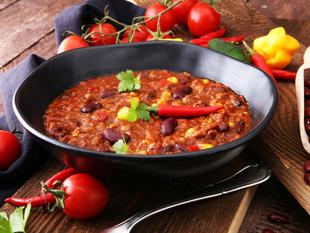 Eine Schüssel mit Chili, garniert mit roten Chilischoten und Koriander, dazu frische Tomaten und rote Kidneybohnen auf einem rustikalen Holztisch.