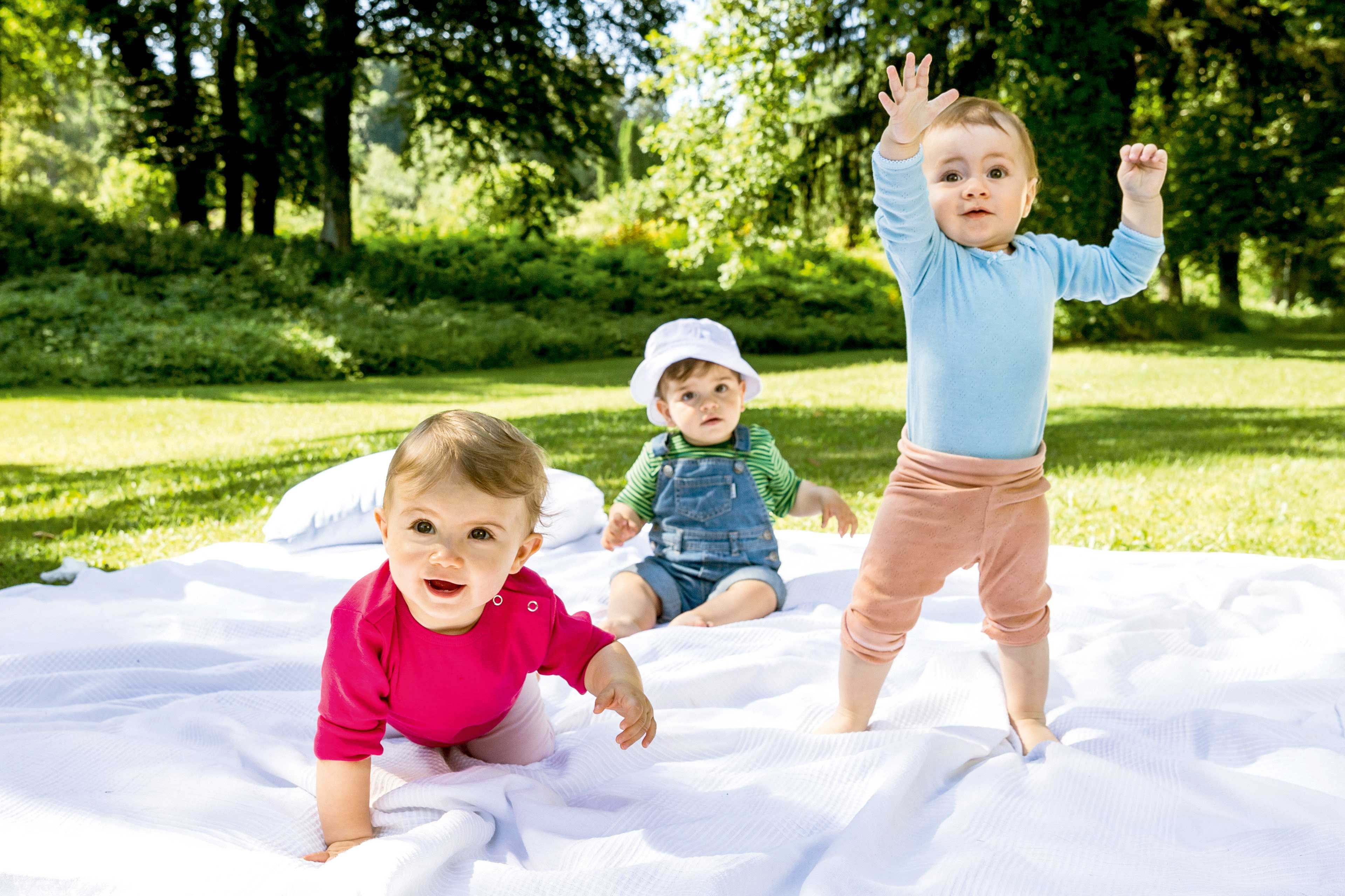 Drei Kleinkinder auf einer weißen Decke im Freien, eines steht mit erhobenen Armen, eines sitzt mit Latzhose und Sonnenhut, eines krabbelt in pinkem Oberteil