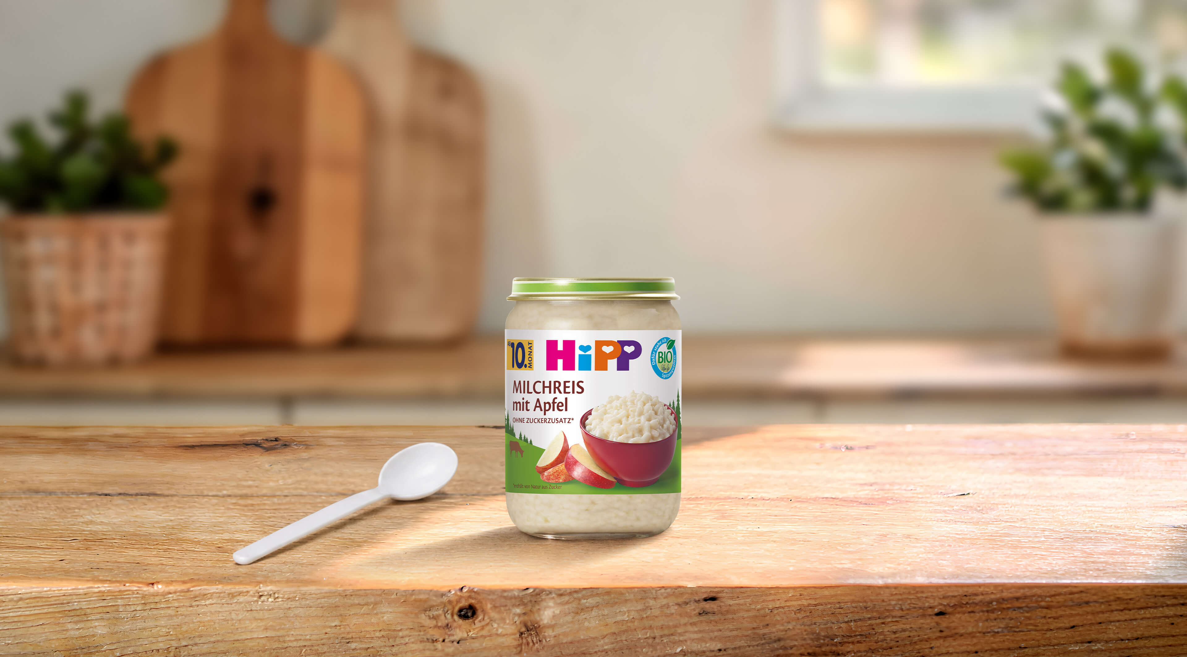 HiPP Bio Milchreis mit Apfel im 190g-Gläschen mit Löffel auf einem Holztisch in einer hellen Küche