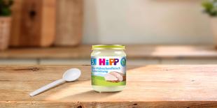 HiPP Bio-Hühnchenfleisch Zubereitung im Glas mit Löffel auf einem Holztisch in heller Küchenumgebung