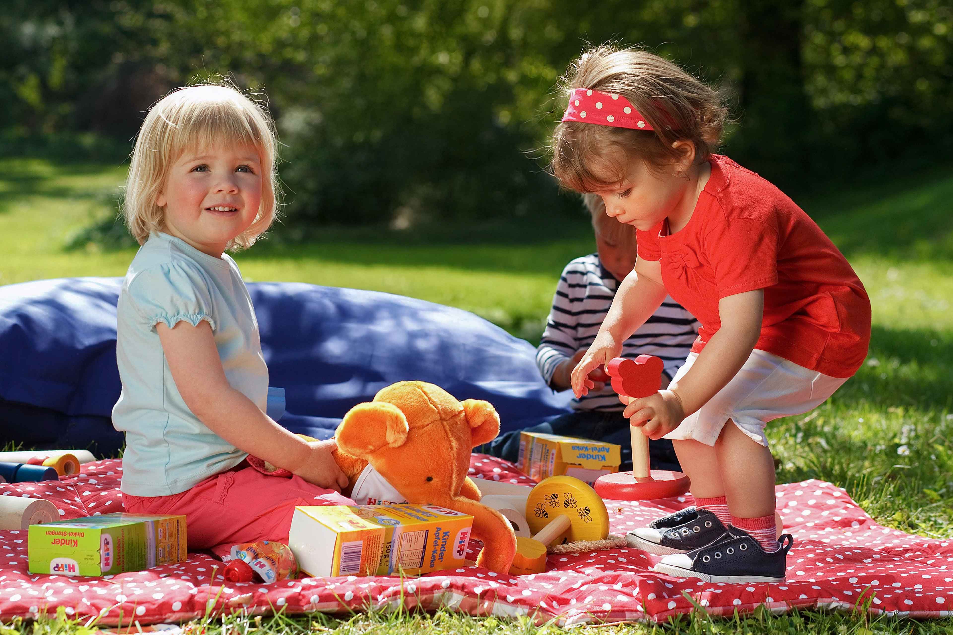 Zwei kleine Kinder spielen im Freien auf einer rot-weißen Decke, umgeben von Spielzeug und einem blauen Kissen.
