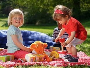 Zwei kleine Kinder spielen im Freien auf einer rot-weißen Decke, umgeben von Spielzeug und einem blauen Kissen.