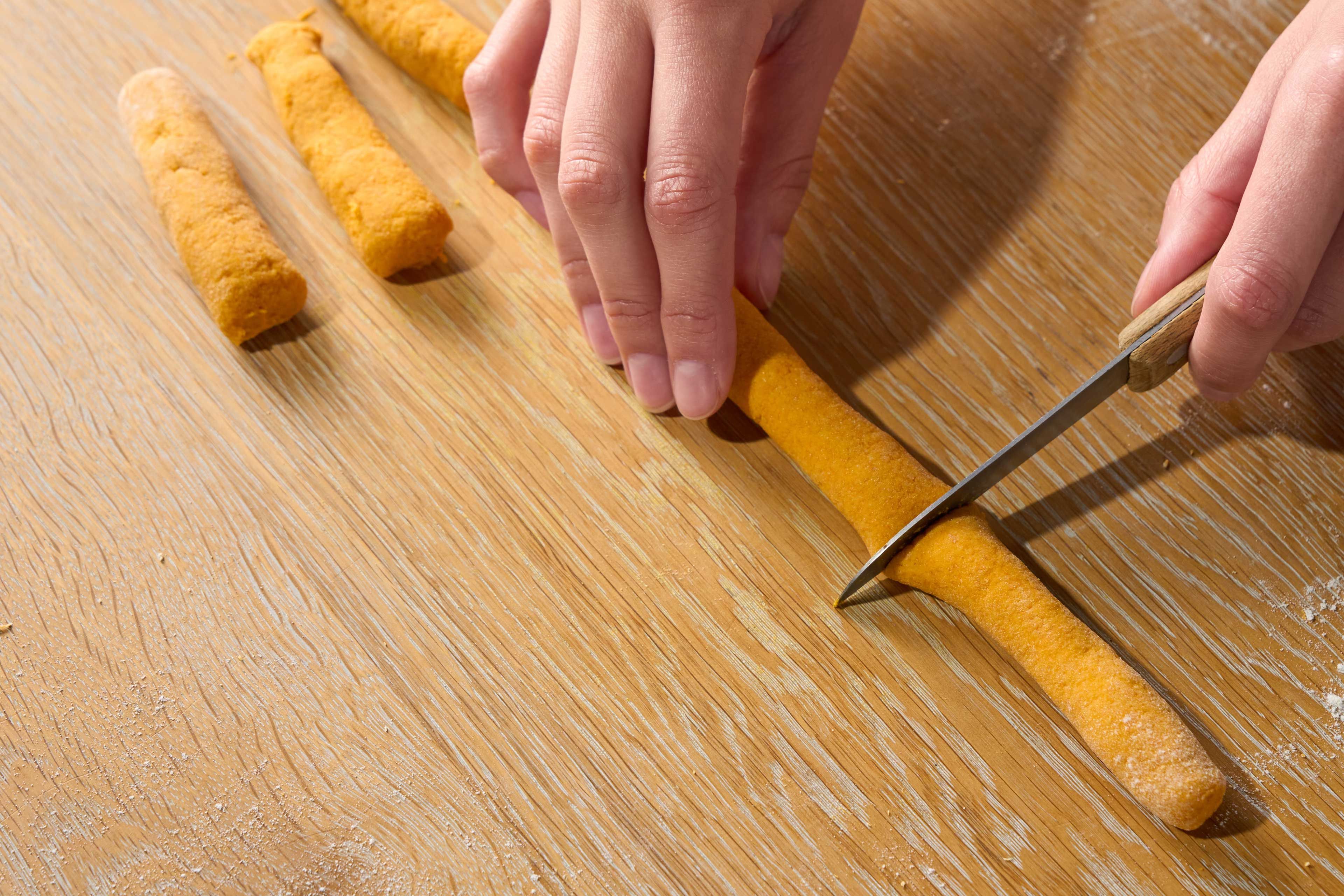 Hände schneiden längliche Teigrollen auf einem Holzbrett in Stücke