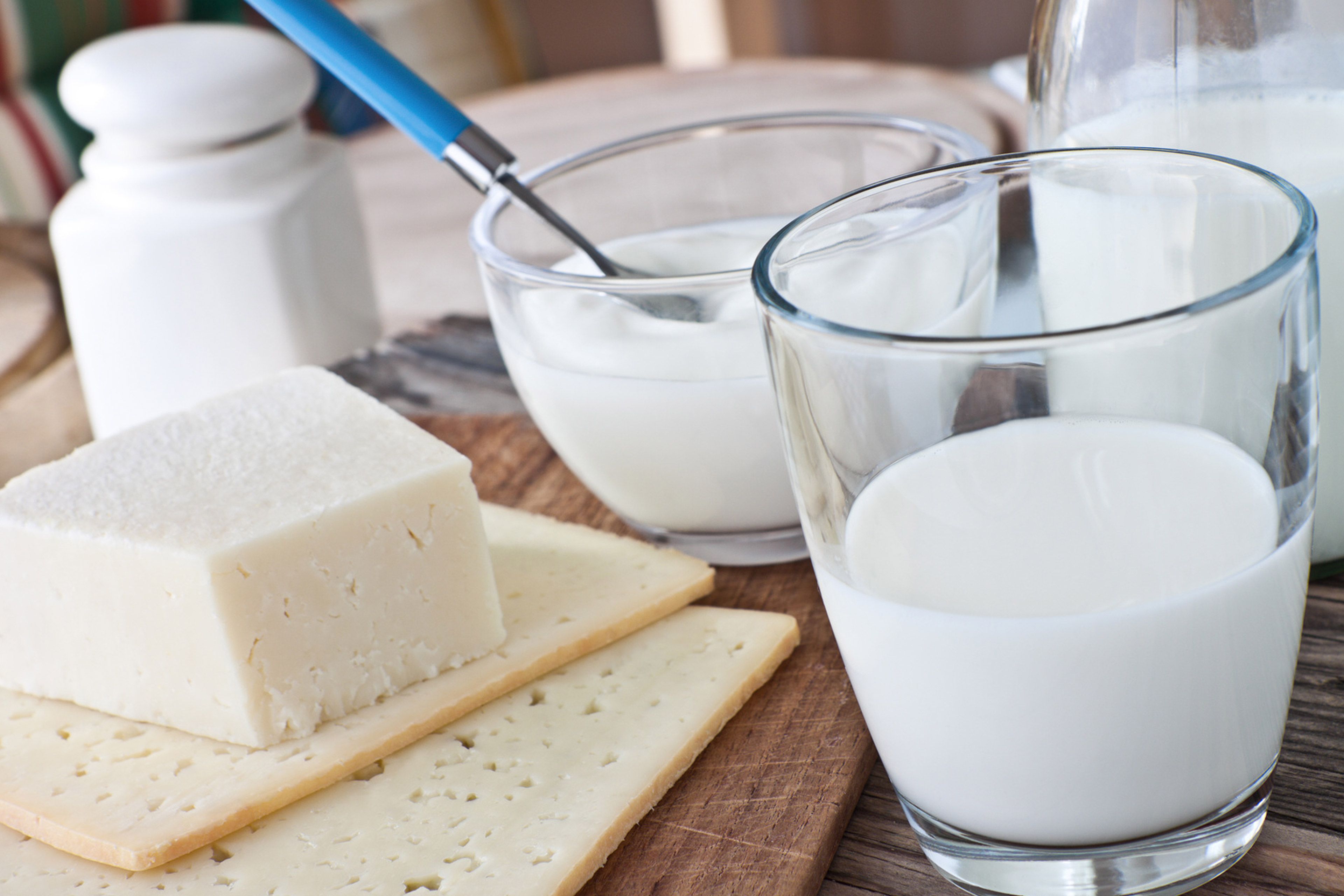 Verschiedene Molkereiprodukte, darunter ein Stück Käse, ein Glas Milch und ein Behälter mit Joghurt, die alle auf einer Holzunterlage platziert sind.