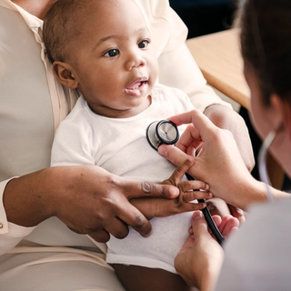 Smiling baby on a parent’s lap having their chest gently checked with a stethoscope during a routine health visit