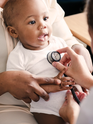 Smiling baby on a parent’s lap having their chest gently checked with a stethoscope during a routine health visit