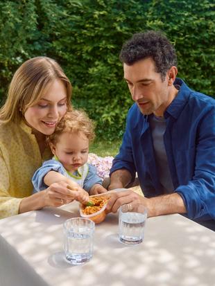 Eltern sitzen draußen im Freien und unterstützen Kleinkind beim Essen von Spaghetti