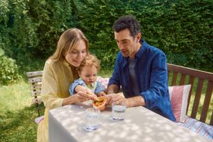 Eltern sitzen draußen im Freien und unterstützen Kleinkind beim Essen von Spaghetti