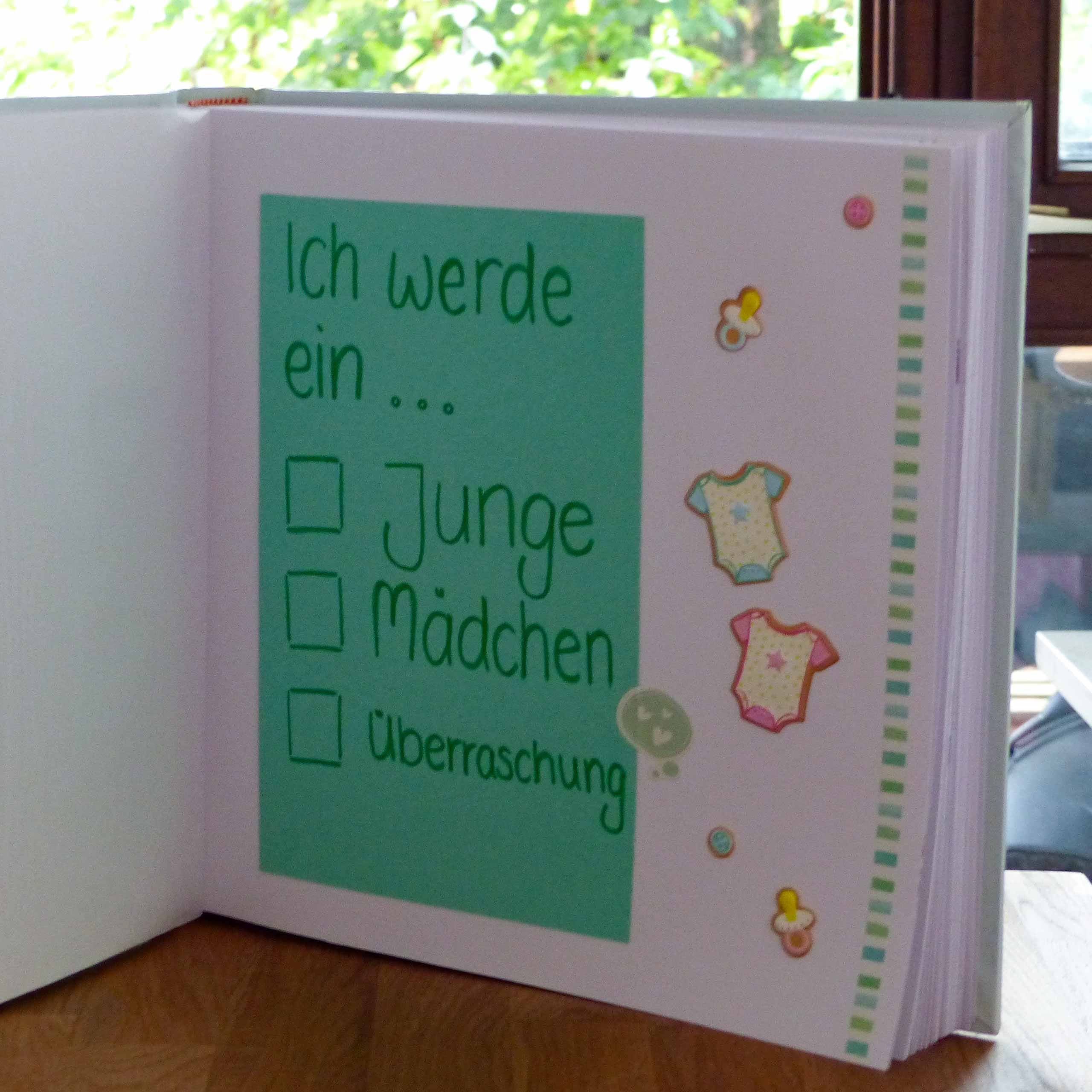 Ein handgefertigtes Buch mit grünem Hintergrund, das eine Liste mit babybezogenen Optionen wie "Junge", "Mädchen" und "Überraschung" enthält, begleitet von dekorativen Aufklebern mit Babykleidung.