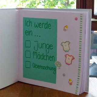 Ein handgefertigtes Buch mit grünem Hintergrund, das eine Liste mit babybezogenen Optionen wie "Junge", "Mädchen" und "Überraschung" enthält, begleitet von dekorativen Aufklebern mit Babykleidung.