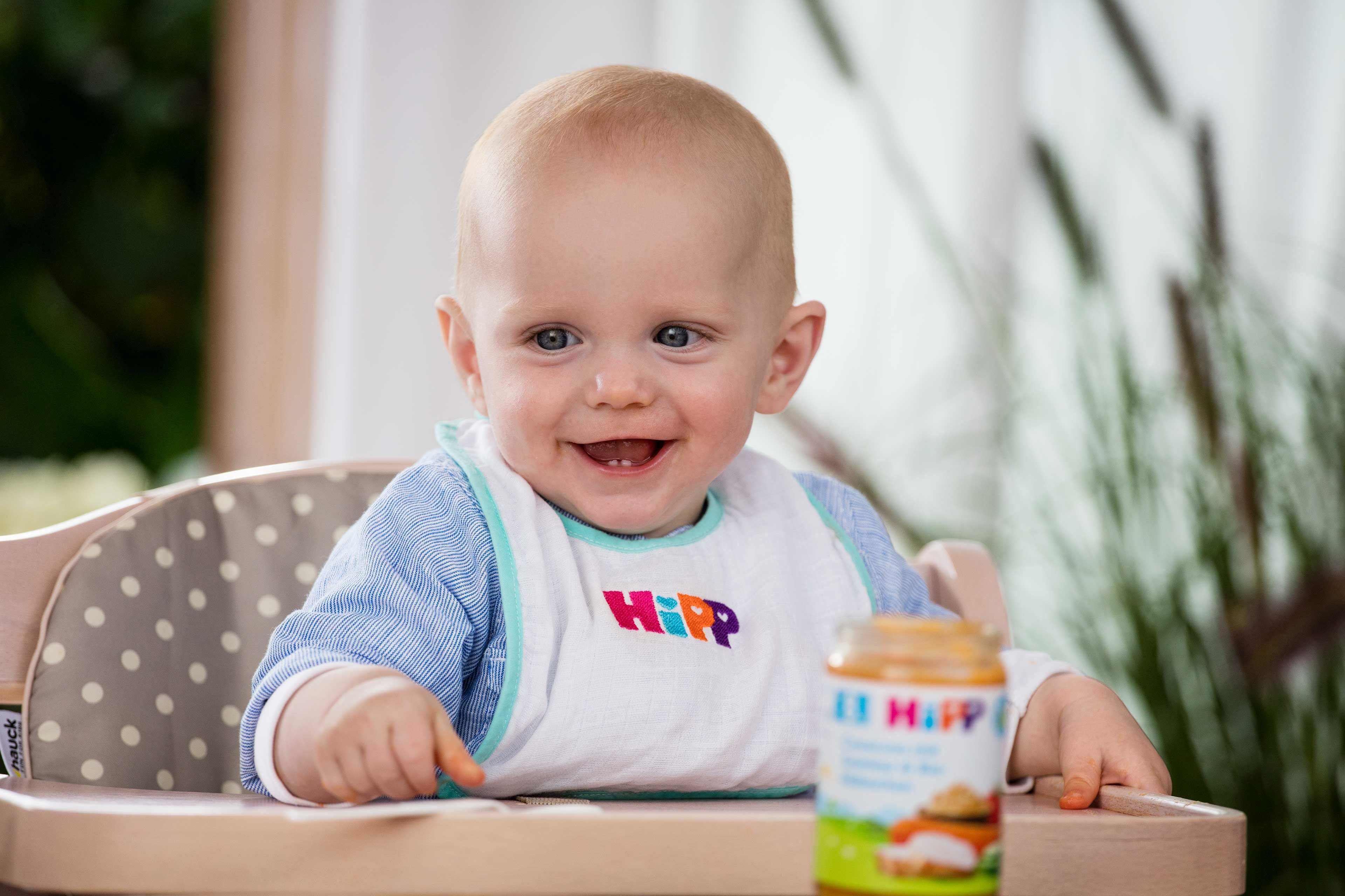 Ein Baby in einem Hochstuhl, das ein Lätzchen mit dem Aufdruck "Hipp" trägt, hält einen Löffel und ein Glas mit Essen in der Hand.