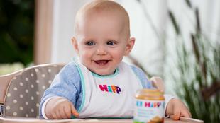 Ein Baby in einem Hochstuhl, das ein Lätzchen mit dem Aufdruck "Hipp" trägt, hält einen Löffel und ein Glas mit Essen in der Hand.