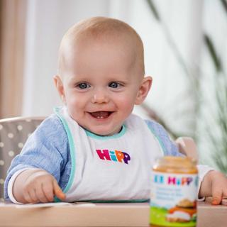 Sitzendes Baby mit blauem Pullover und weißem Lätzchen mit bunter Aufschrift vor einem Glas Babybrei auf einem Tisch
