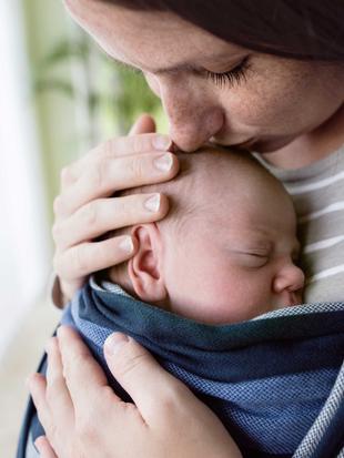 Mutter hält ihr Neugeborenes im Tragetuch dicht an der Brust und küsst liebevoll seinen Kopf für Nähe beim Milcheinschuss