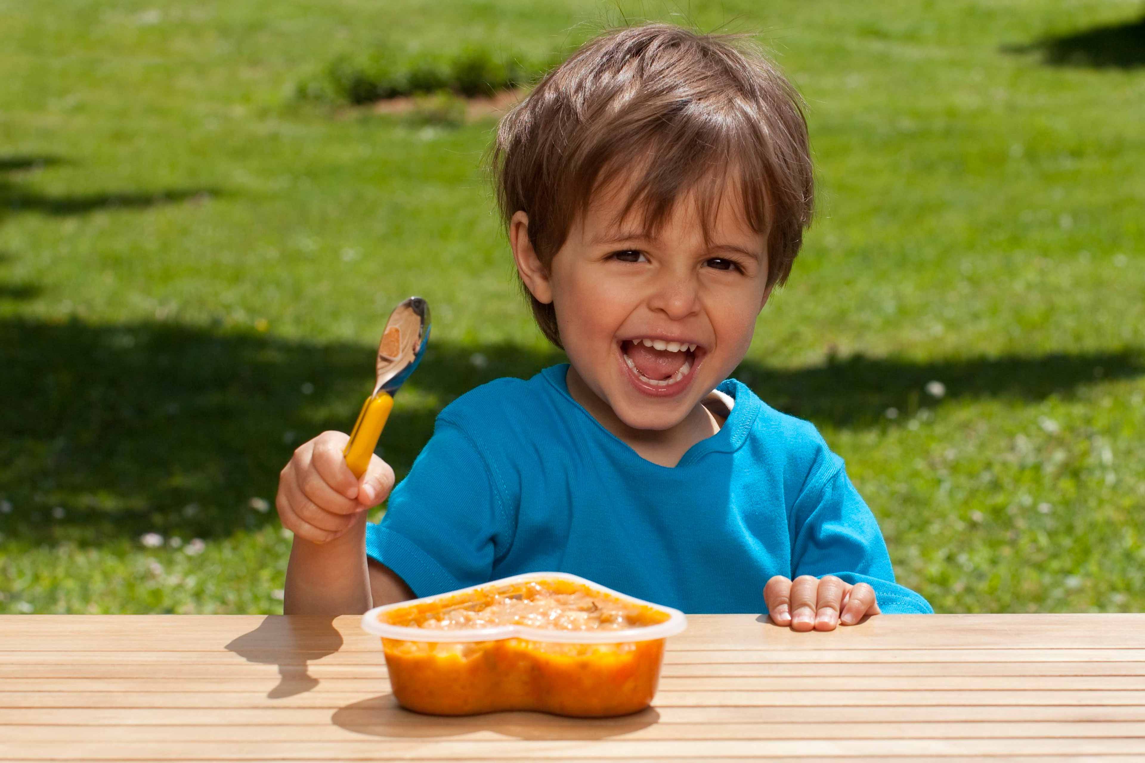 Ein fröhliches Kind in einem blauen Hemd, das einen Löffel und einen Behälter mit orangefarbenem Essen in der Hand hält, vor dem Hintergrund eines grünen Rasens.