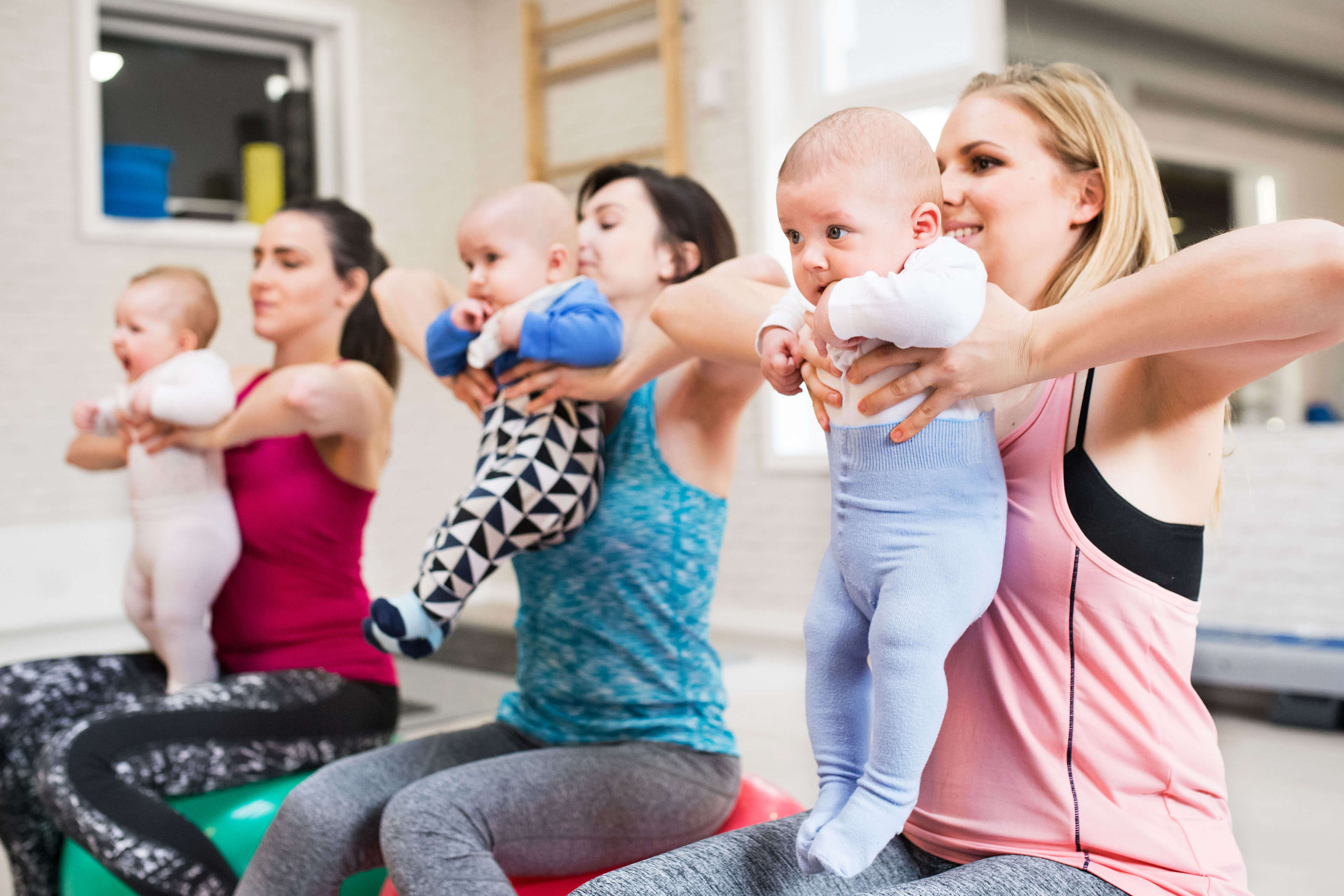 Drei Frauen in einem Fitnessstudio, jede mit einem Baby in der Hand, bei einer Fitnessaktivität.